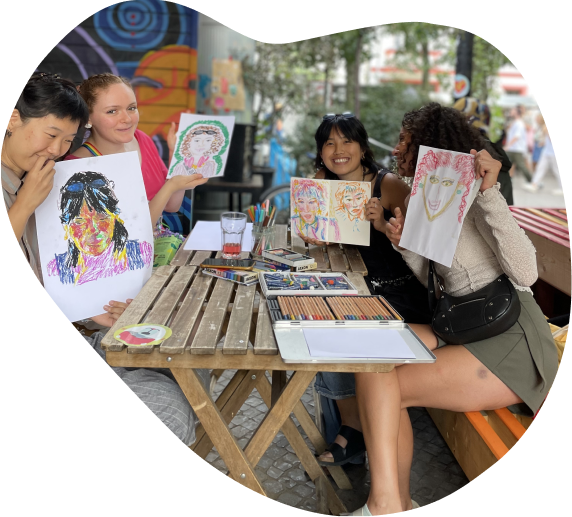 People at an outdoor table holding up colourful self-portraits they have drawn