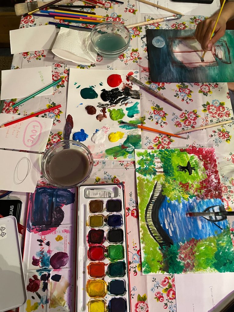 A floral-cloth table covered with paints, brushes, water pots, and colored pencils, with two hands painting landscape scenes during an art workshop.