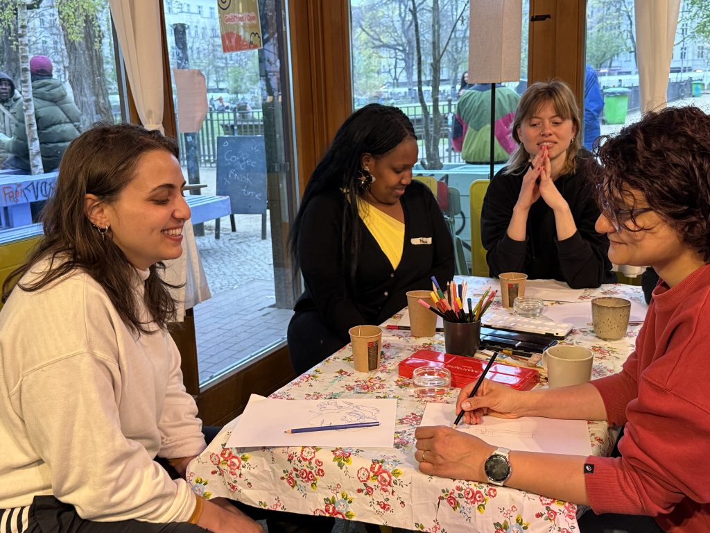 Four women sit at a floral-cloth café table sketching portraits with colored pencils, smiling and chatting, with a park visible through the windows