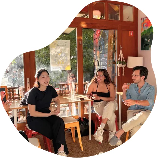 Three people sit and chat over drinks at a sunlit café table, smiling, with large windows and abstract art on the walls behind them